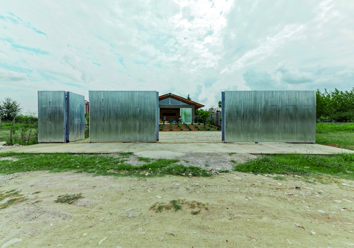 The same corrugated metal gate panels swung open, revealing the villa behind with its gabled roof, concrete platform, and stepped approach