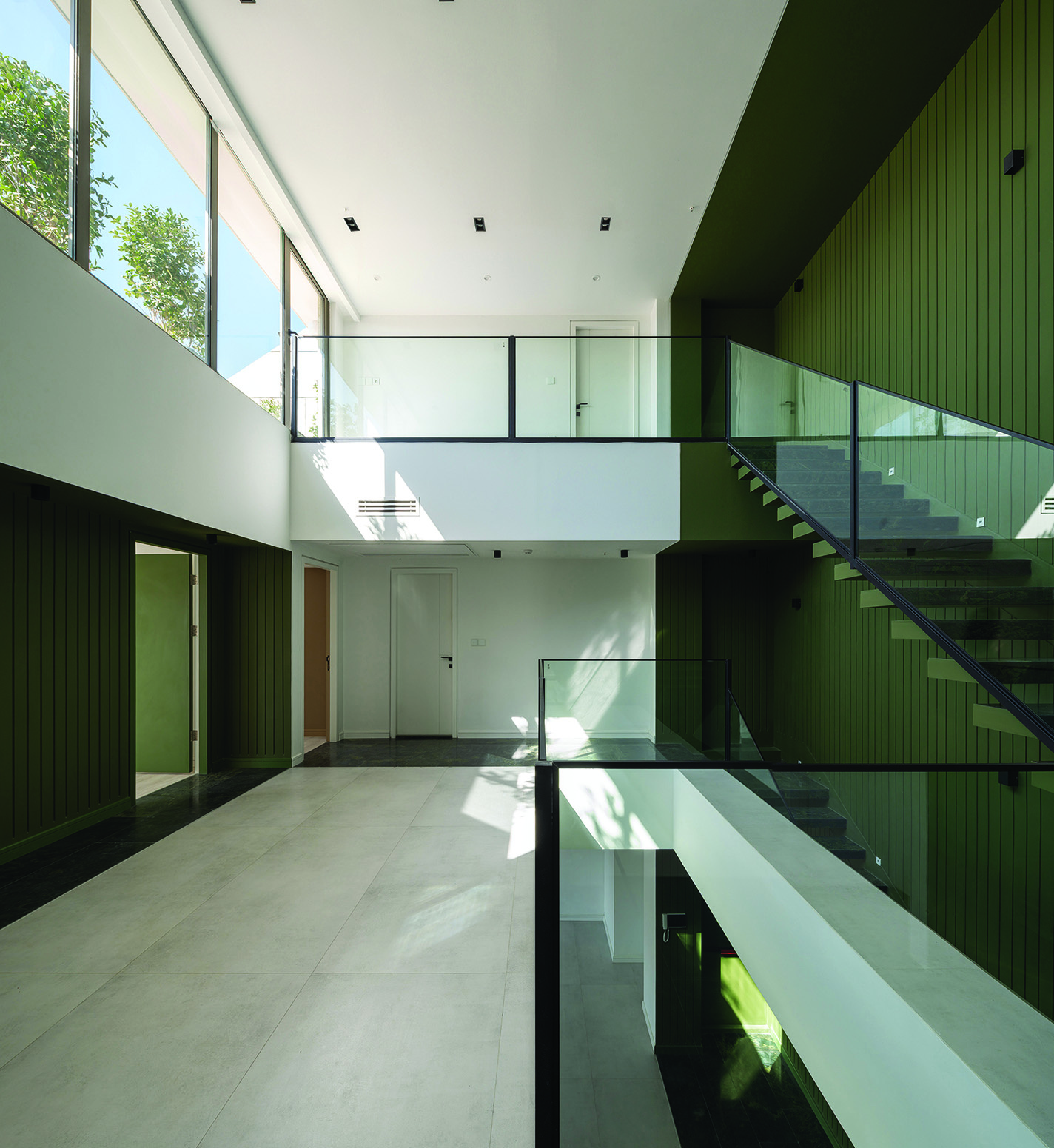Multi-level interior with green-panelled walls, glass railings, floating staircase connecting upper and lower levels, natural light flooding from high windows