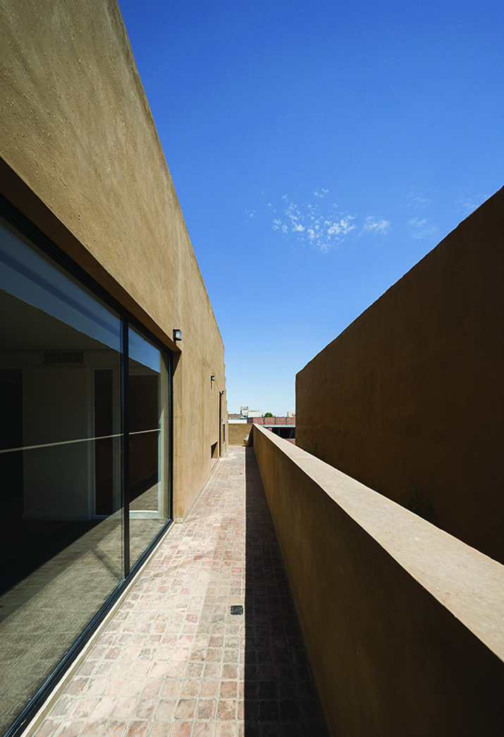 Looking along the narrow passage between two earth-toned walls toward the open sky, brick paving on the floor, a large glass window on the left