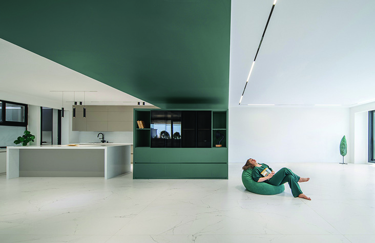 Wide view of the open-plan living and kitchen area with the green volume running across the ceiling, a woman in green clothing lounging on a beanbag on the floor, track lighting overhead, the kitchen island visible to the left