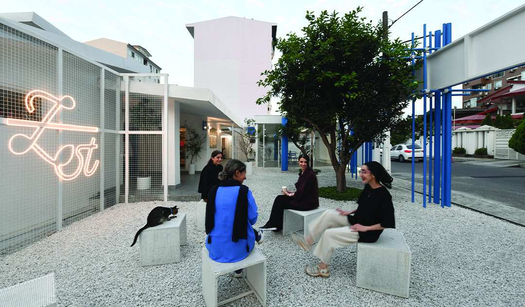 People sitting on concrete cubes in the gravel courtyard at dusk, the Zest neon sign glowing on the mesh facade, a cat resting on one of the cubes, blue scaffolding elements visible