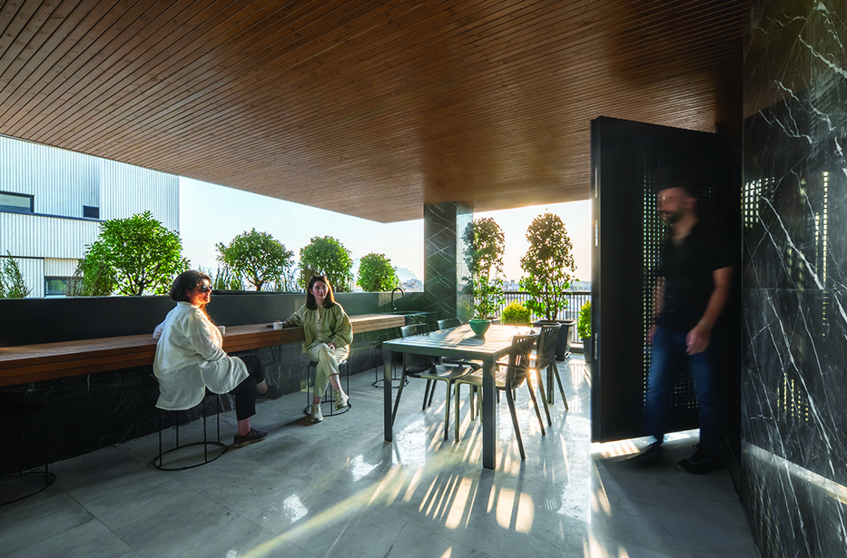 The outer yard at sunset with three people gathered around a table, lush potted trees along the parapet, dark marble walls, warm golden light filtering through the greenery