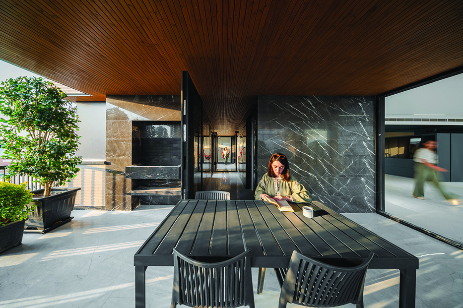 The outer yard terrace with dark marble walls, wood-panelled ceiling, a woman seated at a dining table reading, and a corridor leading into the interior