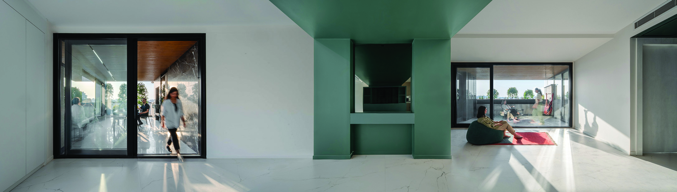 Wide panoramic view of the interior: a woman entering from the terrace through a large glass door on the left, a green-painted volume housing the kitchen and storage in the centre, another woman lounging on a beanbag on a red rug to the right, with a second terrace visible beyond