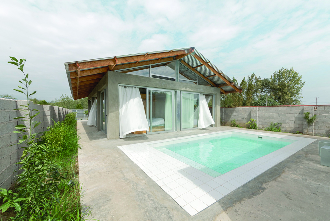 Pati Project from the south in daytime &mdash; concrete villa with gabled roof and white curtains flanking the glass walls, a small pool in the foreground, cinder-block walls