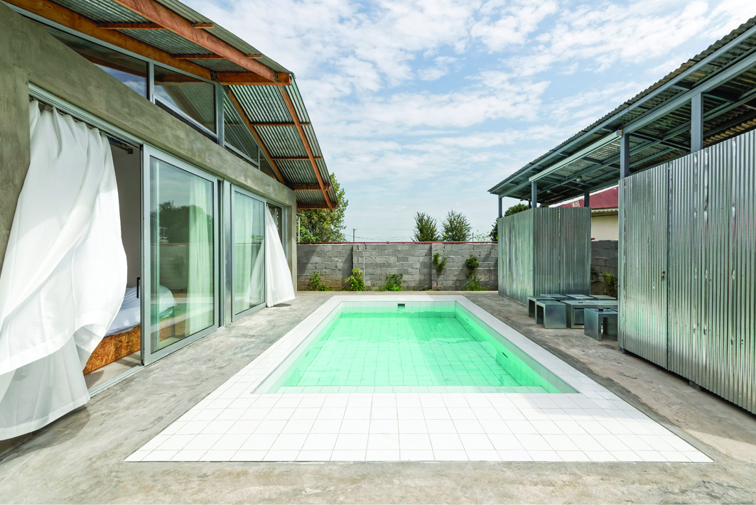 The pool and leisure area of Pati Project &mdash; turquoise pool between the main villa with white curtains on the left and a corrugated metal structure on the right, cinder-block wall in the background