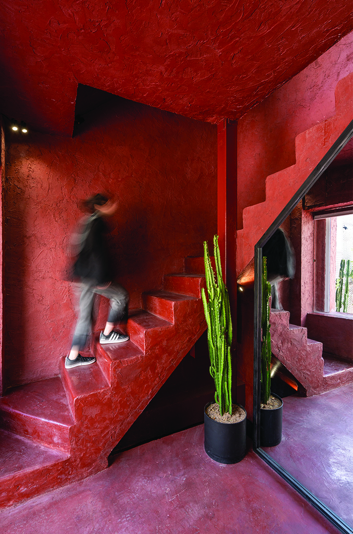 Interior of the red staircase core: a person ascending the red cement stairs, cacti in black pots on the landing, textured red walls and ceiling