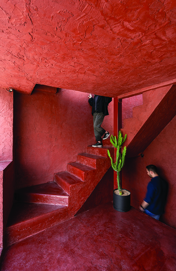 Lower level of the red staircase showing a person descending, textured red cement walls, a cactus in a black pot, and another person seated at the base