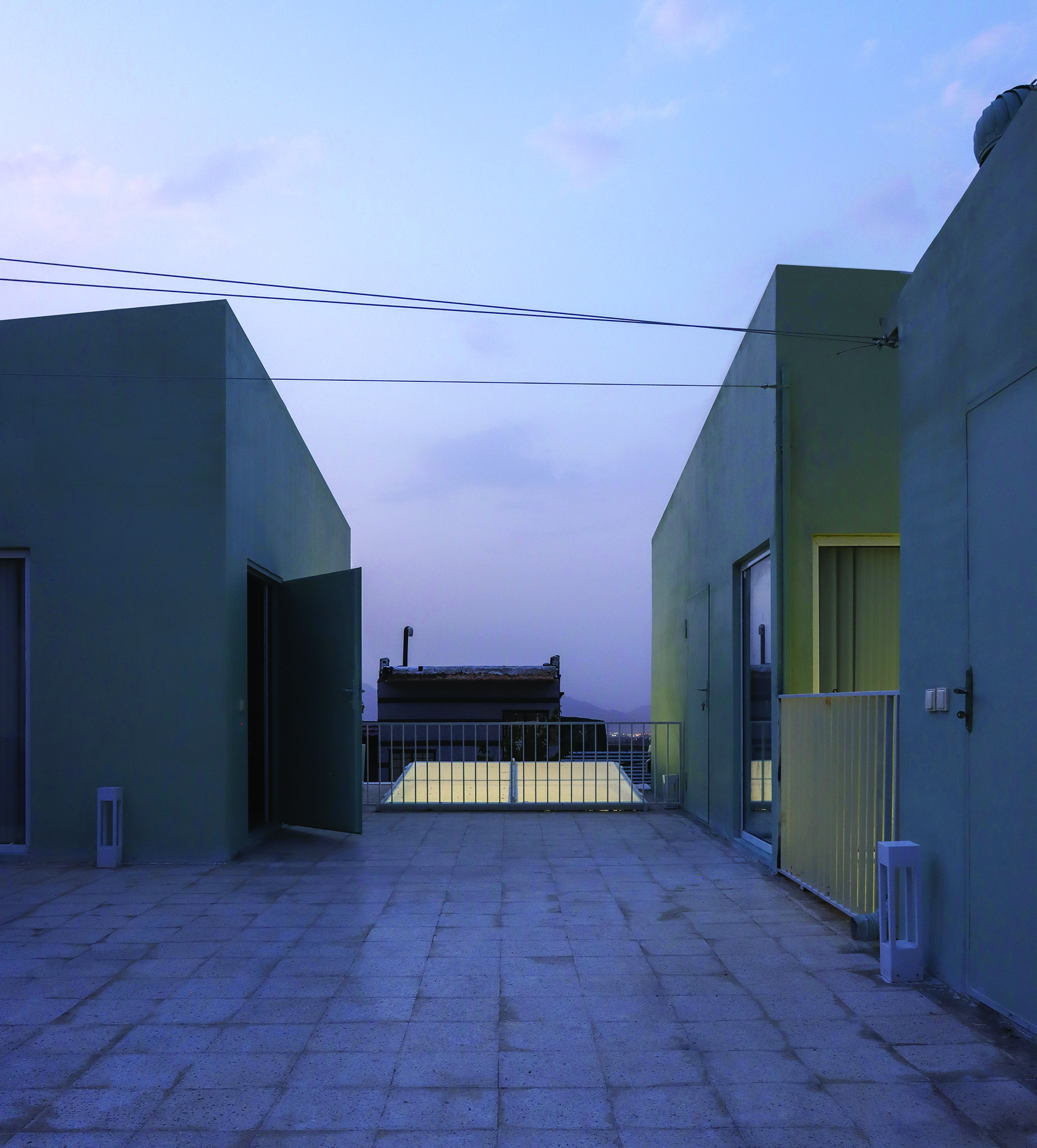 Rooftop courtyard at dusk, looking between two pale green-walled volumes toward the town and mountains beyond, stone pavers on the ground, a warm yellow glow from windows