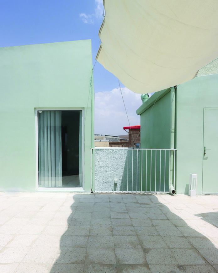 Daytime view of the rooftop terrace with pale green volumes, fabric canopy shade above, white metal railings, a view of red roofs in the distance