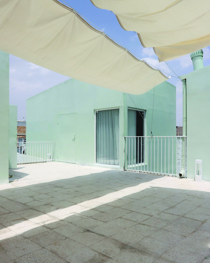 Rooftop terrace under stretched fabric canopy casting shadows on stone pavers, green volumes on either side, blue sky visible above
