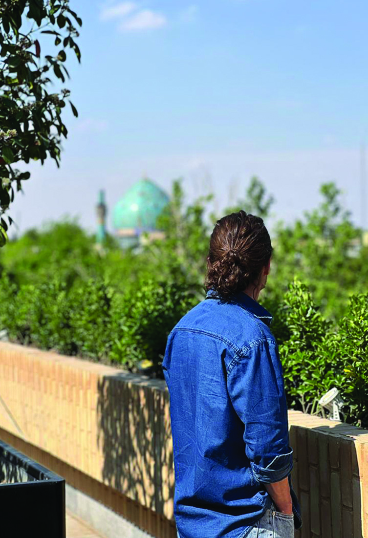 A person standing on the rooftop terrace looking out over the treetops of Chaharbagh toward a turquoise mosque dome in the distance