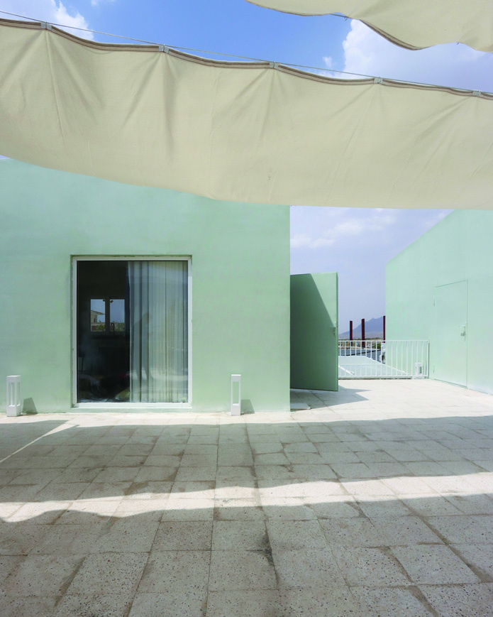 Rooftop view showing a green-painted volume with a large window and curtains, a door open to reveal a vista of the mountains, fabric canopy above casting soft shadows