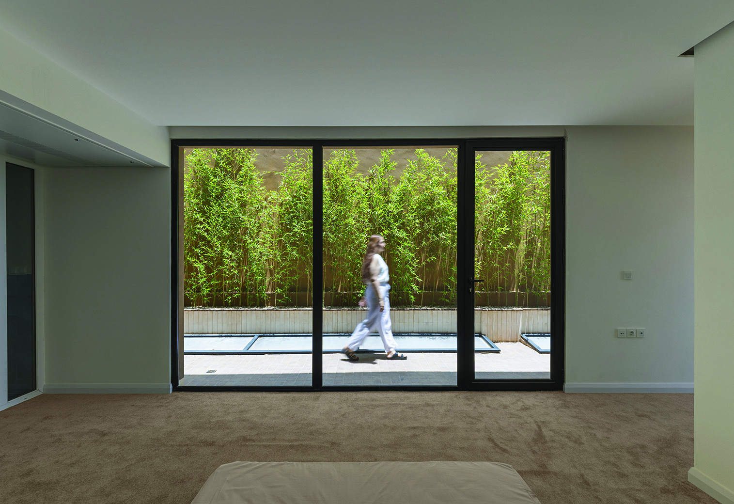 Interior room with large three-panel glass sliding door opening to the side alley, a person walking past bamboo planting outside