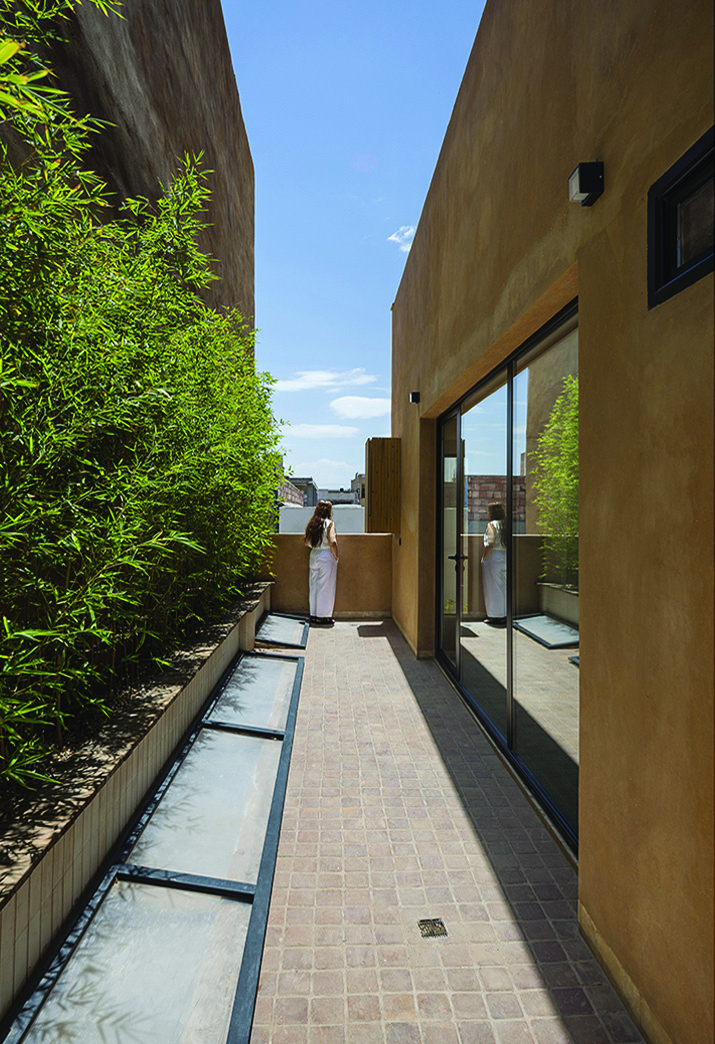 The side alley in daytime &mdash; a woman standing between earth-toned walls with bamboo, glass skylights, and a large glass door to the interior