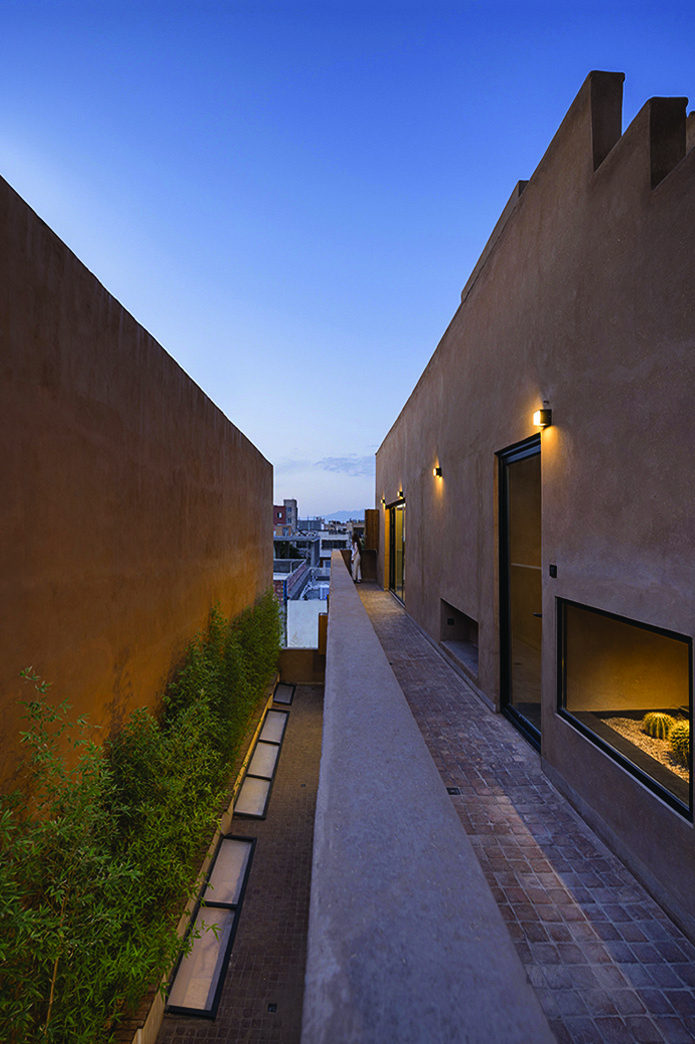 The narrow side alley between Nim Rokh and its neighbour at twilight &mdash; earth-toned walls, bamboo planting, glass skylights in the floor, and warm wall lights