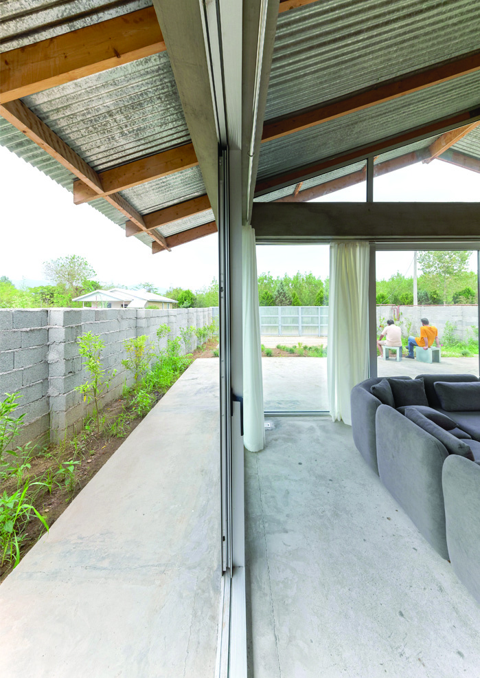 Side corridor of Pati Project showing the open glass wall between inside and outside, corrugated roof overhead, cinder-block wall and vegetation along the side, grey sofa visible inside