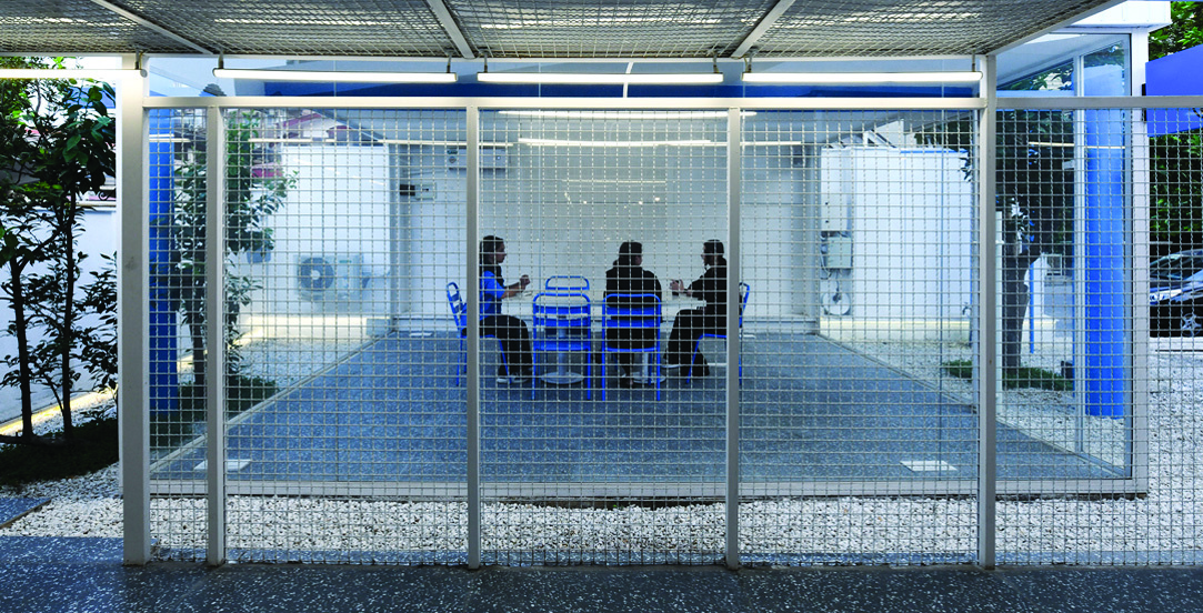 The Social Box: a semi-enclosed space with metal mesh walls and ceiling, people sitting inside on blue chairs at a table, gravel ground, trees visible through the mesh