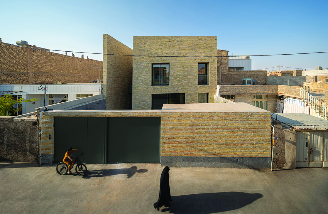 Elevated view of Hamsang House from the south showing the stepped brick volumes, green metal gate, a cyclist and a woman in chador on the lane below