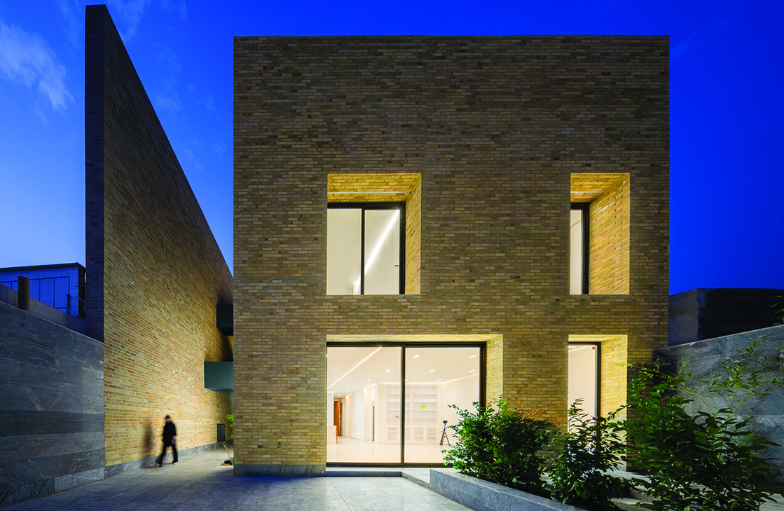 Hamsang House south facade at dusk, brick walls warmly illuminated from within, windows glowing, a person walking through the courtyard