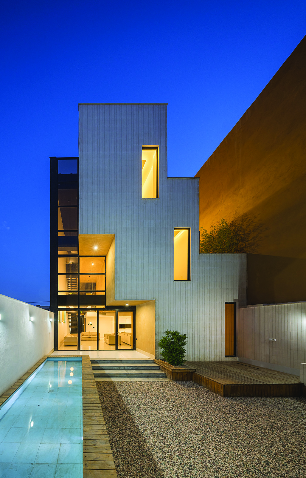 Nim Rokh south facade at twilight &mdash; the stepped building warmly lit from within, with a narrow pool, wooden deck, and gravel courtyard