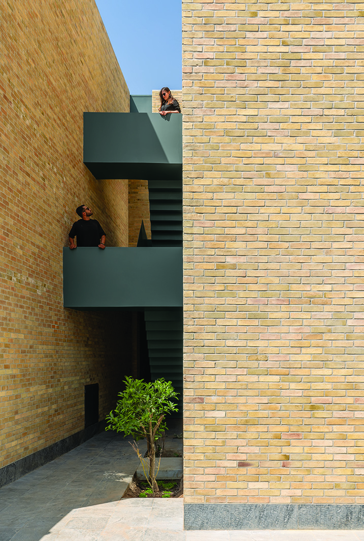 View of the dark green staircase rising between two brick walls, two people on different levels, a small tree growing at the base