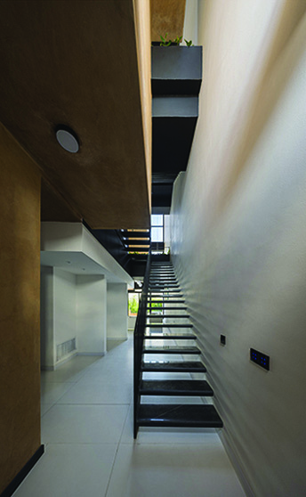 Ground-floor view of the staircase with earth-toned ceiling, white walls, floating steel treads, and daylight filtering from the upper levels