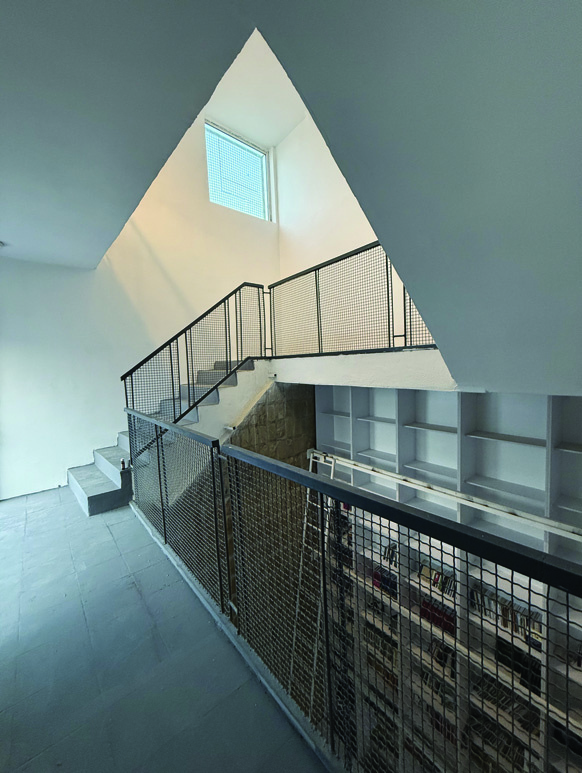 Interior stairwell showing a dramatic triangular skylight above, black mesh railings, concrete stairs, and a view down to the library bookshelves below