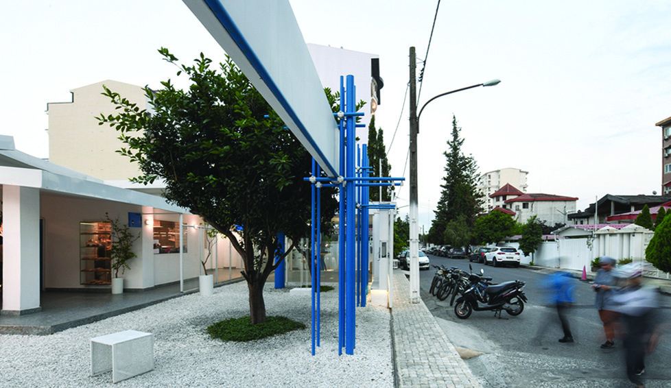 Street-level view showing the cafe entrance with white canopy, blue pipe sculpture, a tree, gravel ground, motorcycles and the neighbourhood street behind