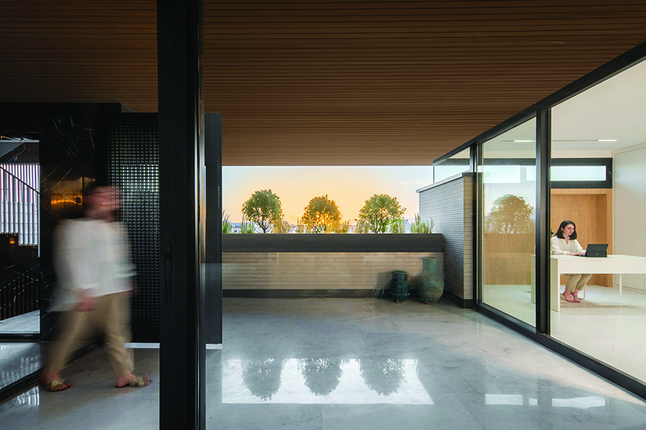 Corridor and terrace at sunset, a blurred figure walking past dark marble walls, a woman working at a desk visible through a large glass window, trees along the parapet catching golden light