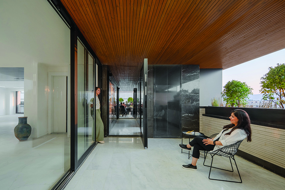Terrace with a woman standing at a glass sliding door and another seated in a wire chair, wood ceiling overhead, greenery along the parapet