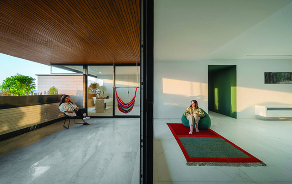 View from the terrace into the living space: a woman seated on the balcony, another lounging on a green beanbag on a red rug inside, a colourful hammock hung between, sunlight streaming in