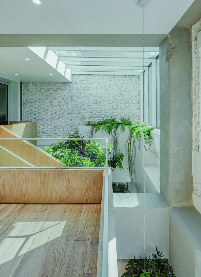 Upper floor gallery with wooden flooring, glass balustrade overlooking the void, trailing plants, exposed concrete column, and skylight above
