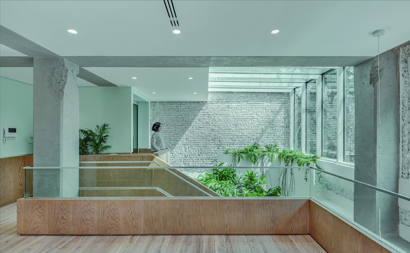A person descending the wooden steps on the upper floor, exposed concrete columns and whitewashed brick wall, lush plants in the void visible through the glass railing