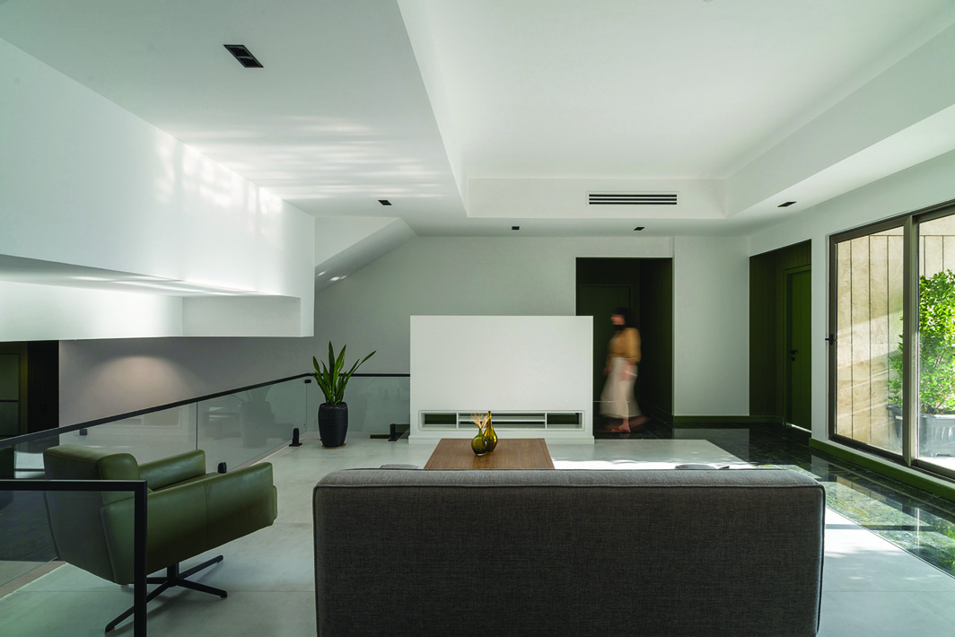 Upper-level living room with grey sofa, green armchair, wooden coffee table, glass railing overlooking lower level, sliding glass door to a terrace with greenery