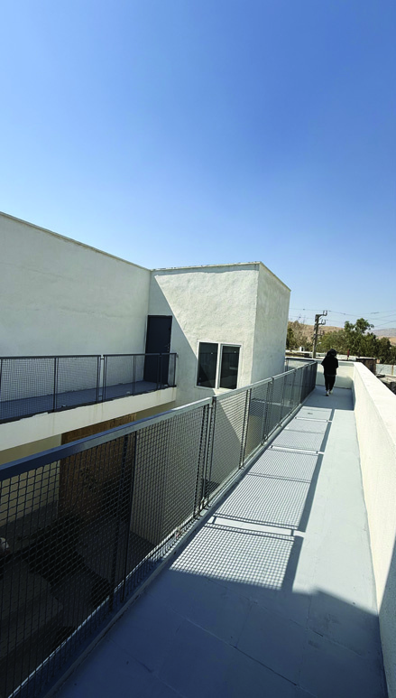 Upper-level walkway with black mesh railings casting geometric shadows, a person walking toward the far end, white concrete walls and blue sky above