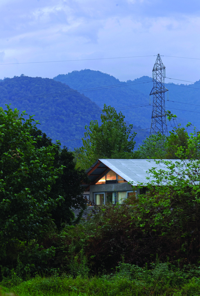 Pati Project seen from a distance at dusk, nestled among green trees with the mountains of Gilan rising behind, a power transmission tower in the background