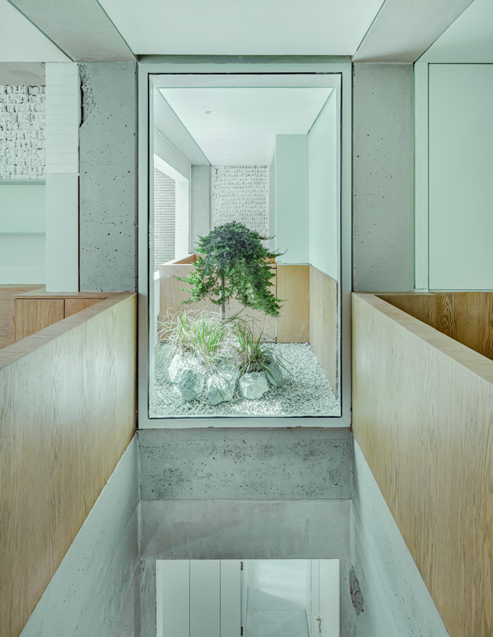Looking down into the central void from the upper floor, with a tree growing up through the opening, white pebbles and rocks at its base, concrete walls and wooden balustrades framing the view