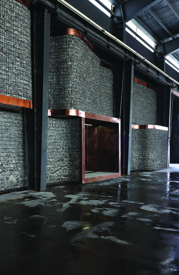 The habitable wall seen from the showroom side &mdash; tall gabion panels with copper-framed window openings between them, wet concrete floor reflecting the forms