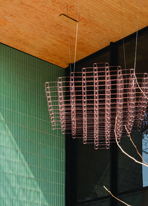 A wire-mesh sculpture suspended from the wooden ceiling of the portico against a green-tiled wall and tree branches