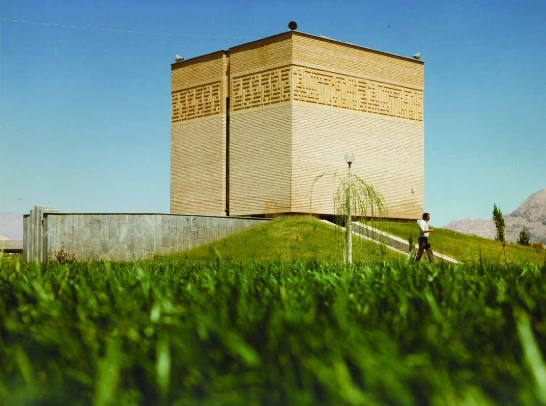 Prayer hall exterior, a cube-shaped brick building on a green hill with mountains in background