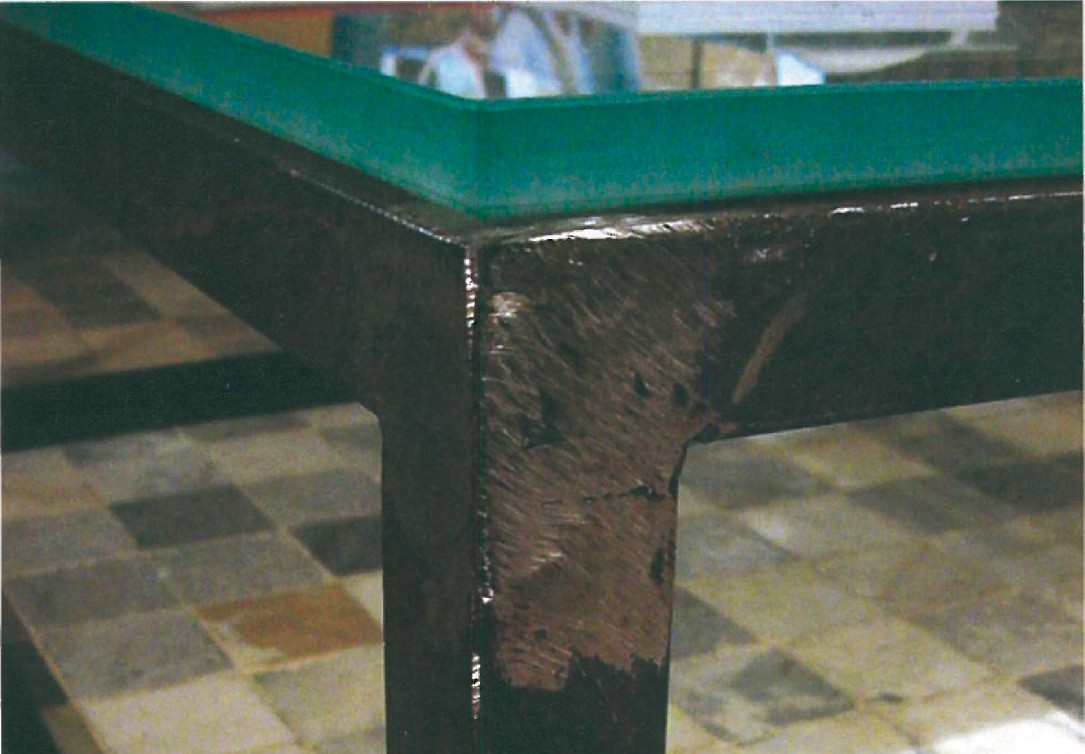 Detail view of the L-frame metal coffee table with thick sand-blasted glass top resting on a stone-tile floor