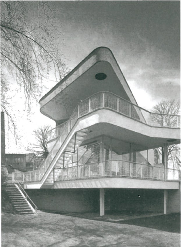 B&W photograph of Hans Scharoun's Haus Schminke — the curved cantilevered upper deck floating over a glass-and-steel pavilion in a wooded suburban garden, captured looking up at the south elevation.