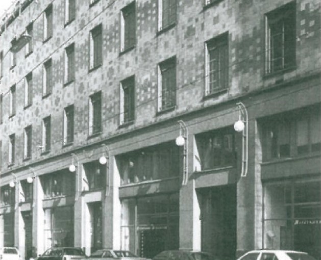 B&W photograph of Adolf Loos's Goldman & Salatsch building (Looshaus) in Vienna (1909-1911) — long brick facade with arched ground-floor shop bays and globular street lamps, parked cars along the kerb.