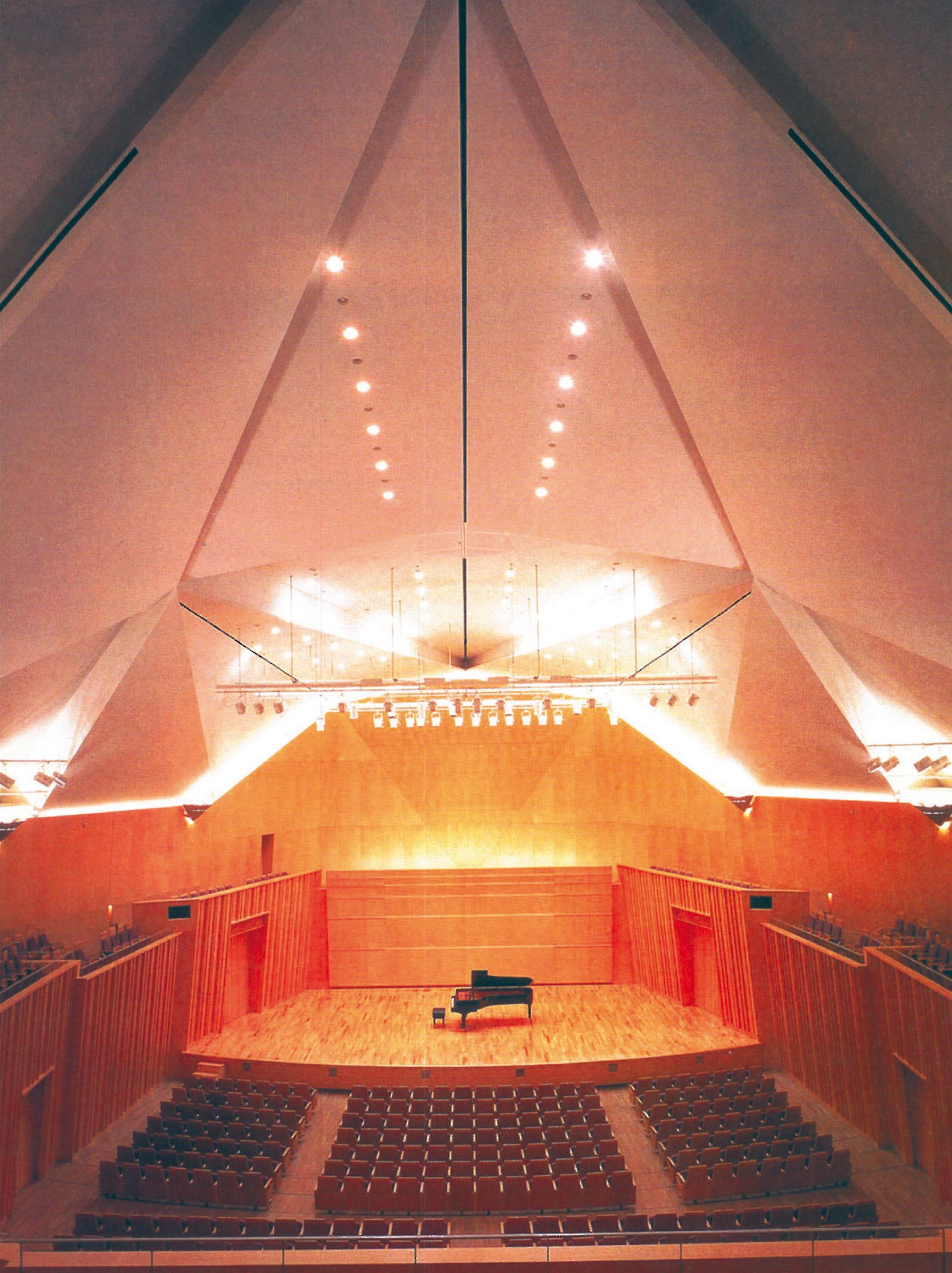 Full-page interior view of the Kirishima/Iwojima concert hall by Maki — triangular folded ceiling, downlights, raked auditorium seating and a single grand piano on the stage