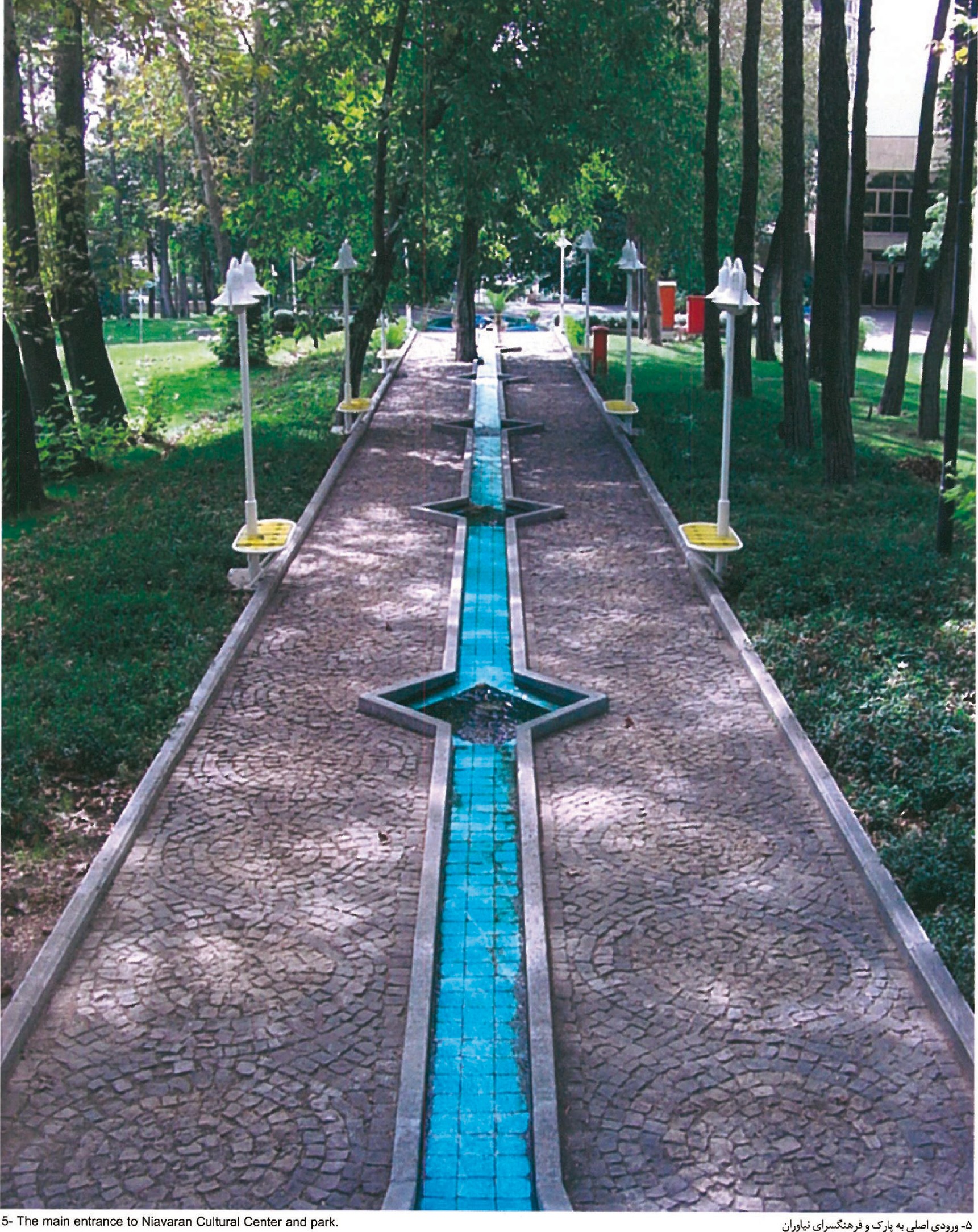 View along the main entrance pathway to the Niavaran Cultural Center — a narrow blue-tiled water channel runs down the centre of a stone-paved walk between rows of new lamp standards under mature trees.