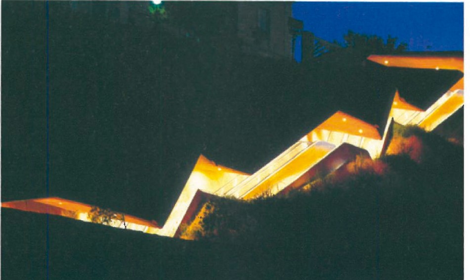 Night view of the same Toledo urban escalator under warm lighting — the zigzag form glowing against the hillside