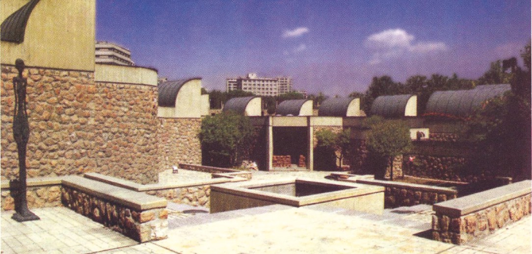 Aerial photograph of the Tehran Museum of Contemporary Arts by Kamran Diba — its row of curved skylit gallery vaults set behind a low rubble-stone wall, with a Giacometti sculpture on a plinth in the courtyard.