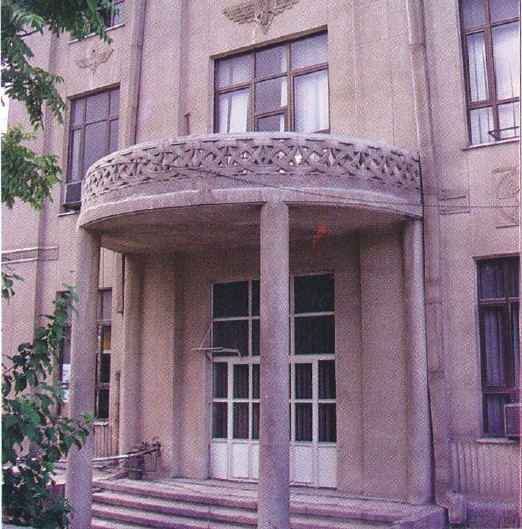 Photograph of Tehran's main Railway Station — limestone-clad neoclassical entrance with the winged emblem of Iran above the canopy, double doors and stone balustrade carved with stylised ornament.