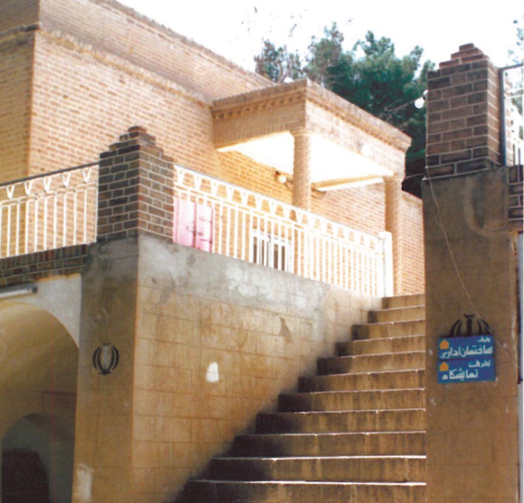 Akbarieh garden building in Birjand: a brick gateway with broad concrete stairs ascending to an open terrace, an Iranian flag detail and a blue notice board for the administration office